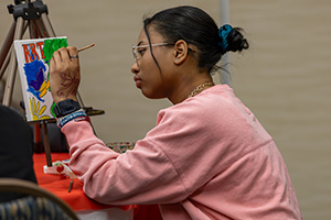 A person in a pink sweatshirt painting on a small canvas on an easel.