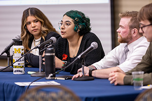 A panel discussion with four speakers seated at a table with microphones, water bottles, and a notepad in front of them.