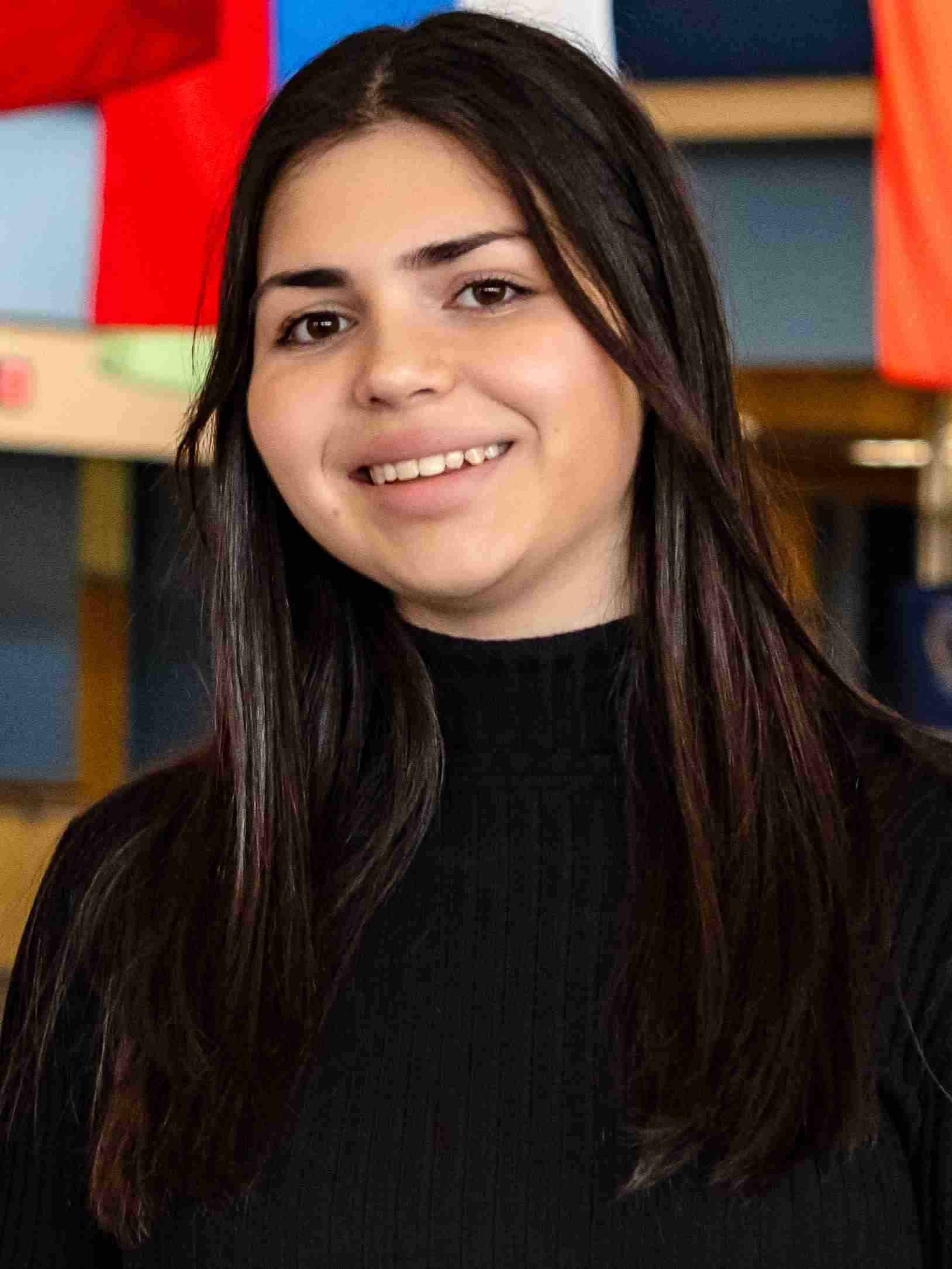 A headshot of Angela Qazaha, a young woman with long, straight dark hair. She is smiling with her teeth visible, wearing a black ribbed turtleneck against a brightly colored background that includes portions of international flags.