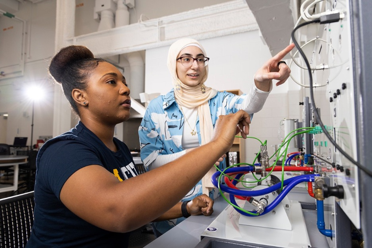 Two individuals are working together on a technical setup, with one pointing at equipment while the other assists.