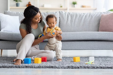 A caregiver and a child are playing on a gray rug with colorful toys scattered around them.