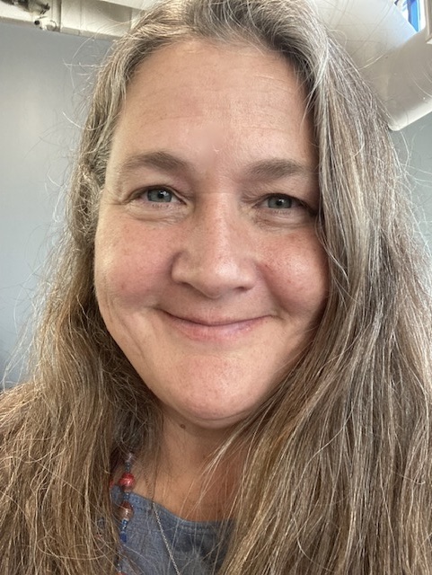 A person with long, wavy hair wearing a gray shirt and a necklace, sitting indoors.