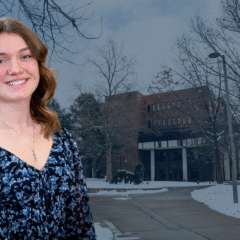 A woman with wavy hair wearing a floral blouse stands in front of a snow-covered campus building.
