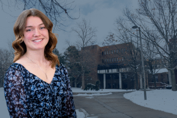 A woman with wavy hair wearing a floral blouse stands in front of a snow-covered campus building.