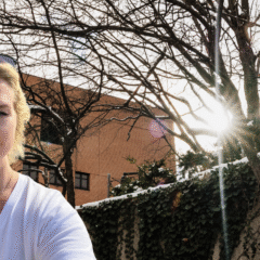 A person wearing a white shirt stands outdoors with bare trees and sunlight shining through them, against a building backdrop.