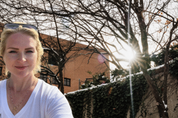 A person wearing a white shirt stands outdoors with bare trees and sunlight shining through them, against a building backdrop.