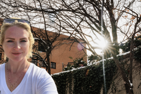 A person wearing a white shirt stands outdoors with bare trees and sunlight shining through them, against a building backdrop.