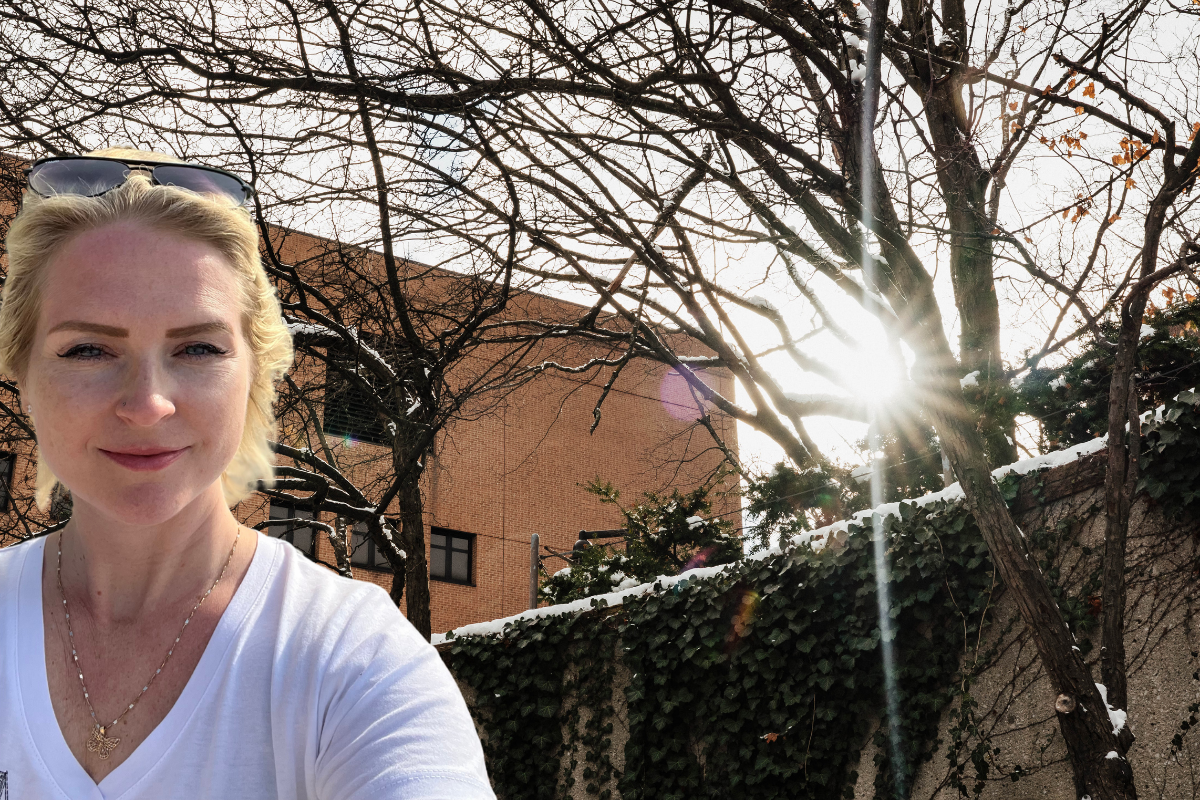 A person wearing a white shirt stands outdoors with bare trees and sunlight shining through them, against a building backdrop.