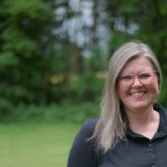 A woman with long, straight, light brown hair wearing a black shirt stands outdoors in a green, wooded area.
