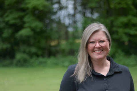 A woman with long, straight, light brown hair wearing a black shirt stands outdoors in a green, wooded area.