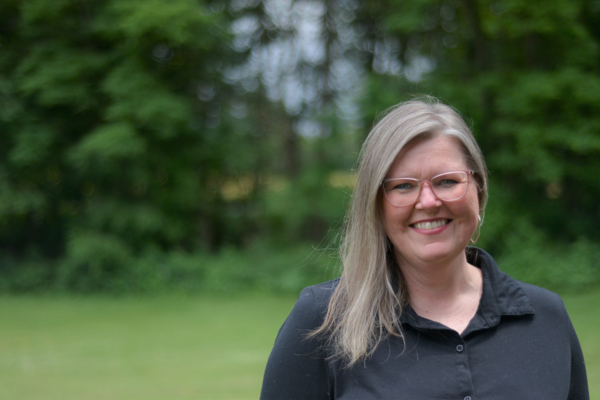 A woman with long, straight, light brown hair wearing a black shirt stands outdoors in a green, wooded area.