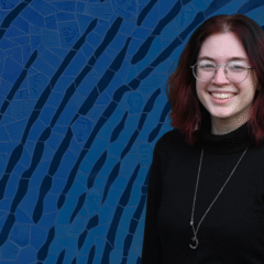 A person stands in front of a blue mosaic background featuring various faces and text inscriptions.