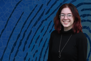 A person stands in front of a blue mosaic background featuring various faces and text inscriptions.