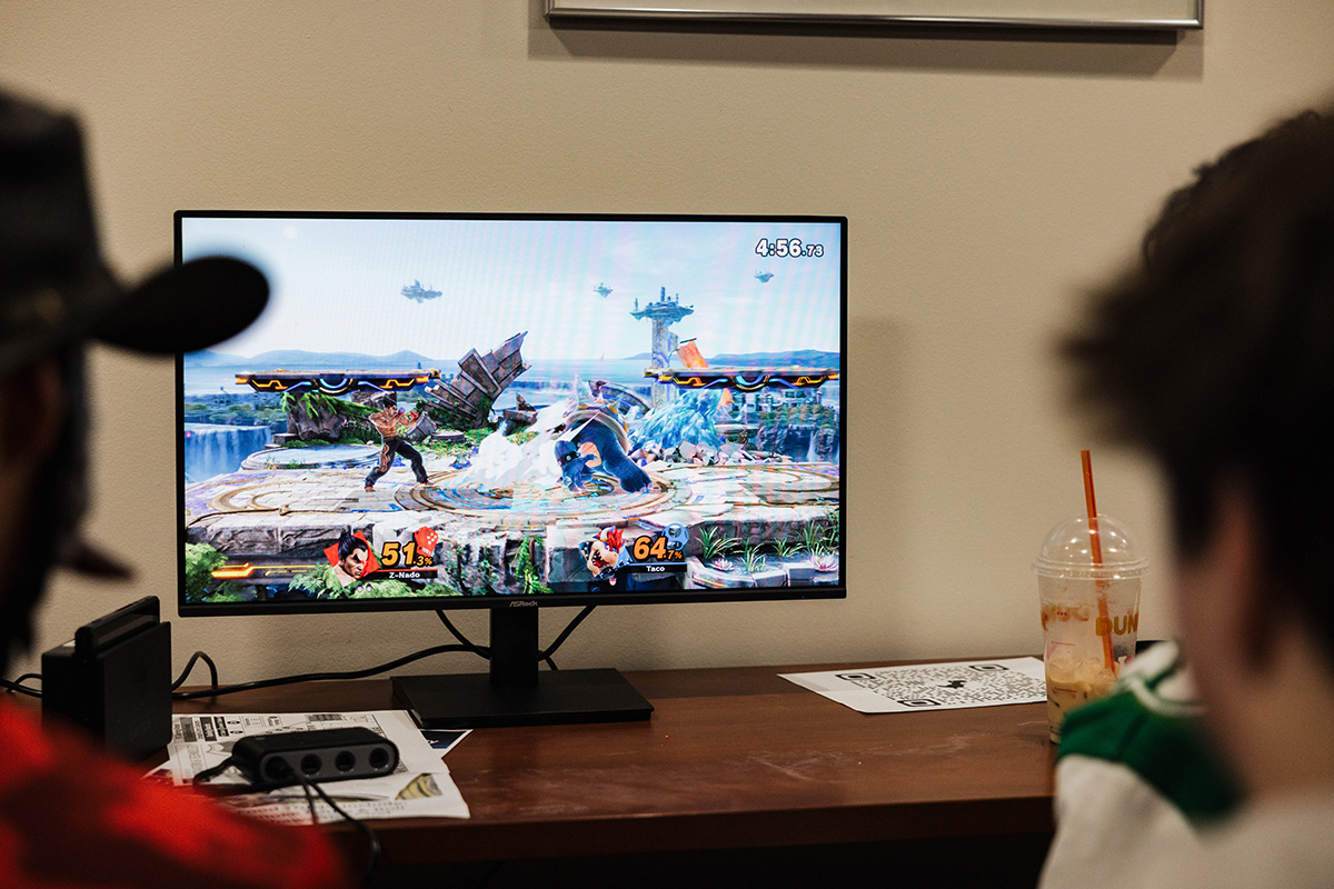 A gaming monitor displays a fighting game with two characters battling. One character has 51.3% damage and is labeled "Z-Nado," while the other has 64.7% damage and is labeled "Taco." A timer shows 4:56 remaining. In the foreground, a person is partially visible, and a drink is on the table.