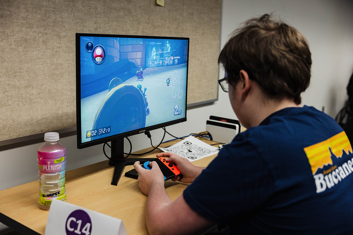 A person with short hair, wearing a blue shirt, is playing a racing video game on a monitor. The screen shows a colorful game scene with characters and a track. A bottle of flavored water is on the table, and a sign labeled "C14" is visible.