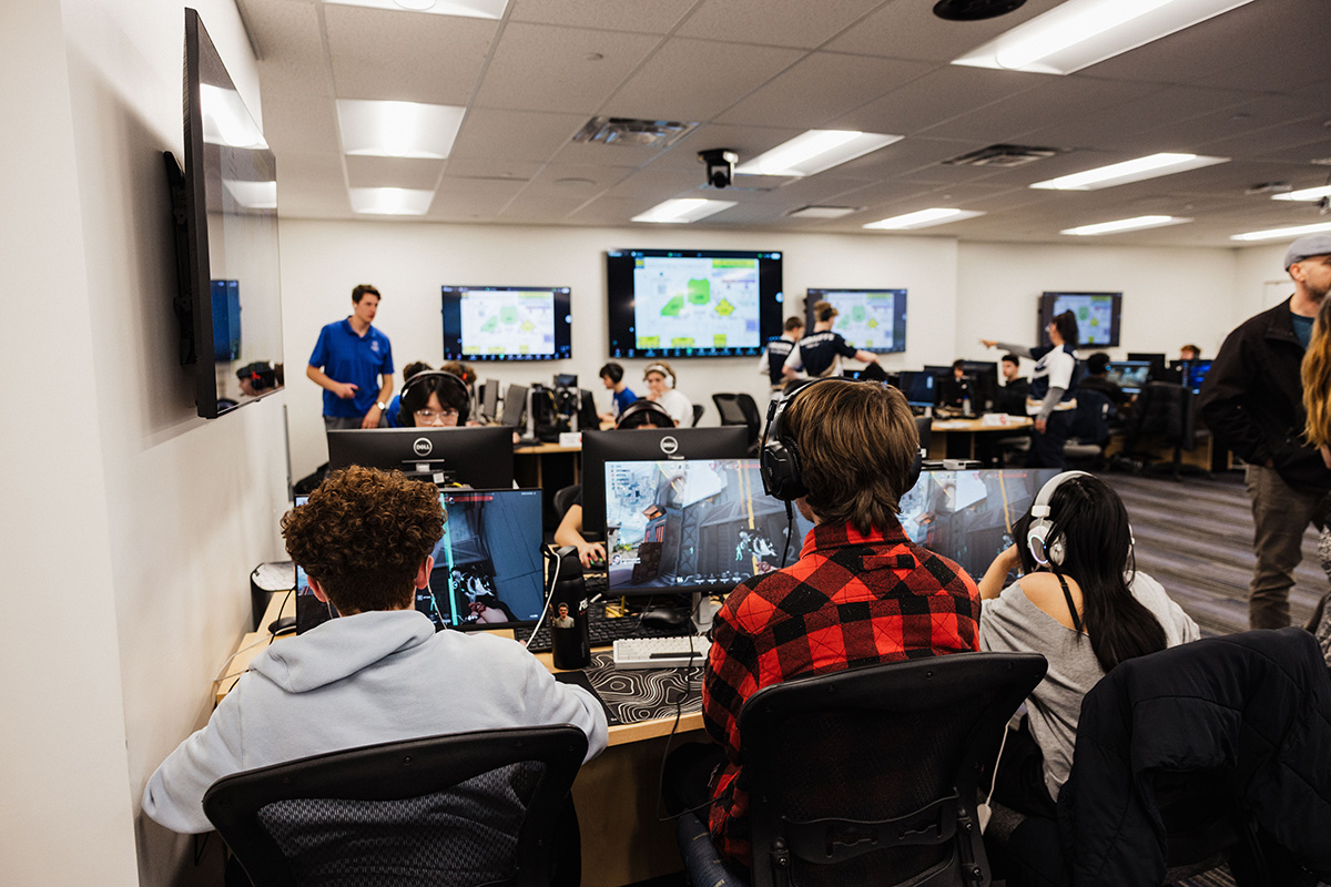 A group of people in a computer lab, focused on gaming, with multiple screens displaying game graphics and maps.