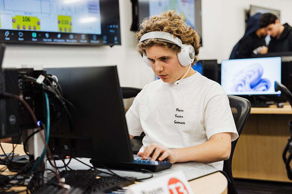 A person wearing headphones is sitting at a computer, typing on a keyboard. Multiple screens are visible in the background displaying various information. A sign on the table indicates "Room 13."