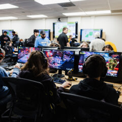 A group of people is gathered in a gaming room, with several individuals seated at computers wearing headphones. The screens display colorful video game action, while others are standing and interacting in the background.