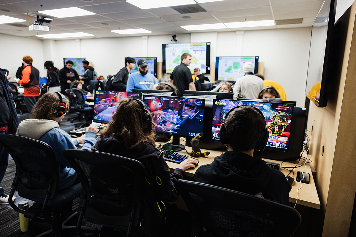 A group of people is gathered in a gaming room, with several individuals seated at computers wearing headphones. The screens display colorful video game action, while others are standing and interacting in the background.