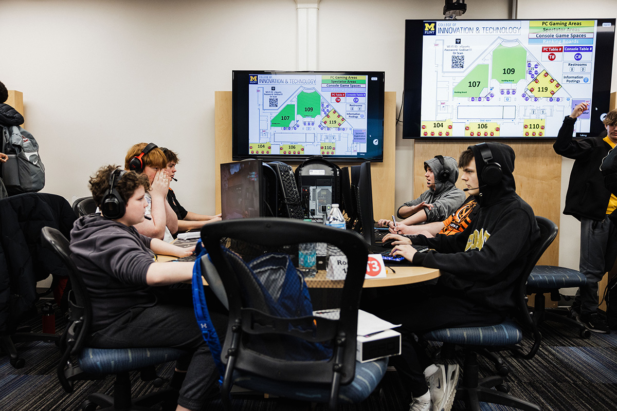 A group of individuals is seated at computers in a gaming area, wearing headphones. Two large screens display a map of the facility and information about gaming spaces.