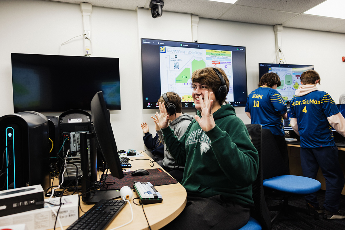 A computer lab with multiple monitors displaying a map and information. One person is sitting at a desk with headphones, raising their hands, while others are standing in the background wearing jerseys.