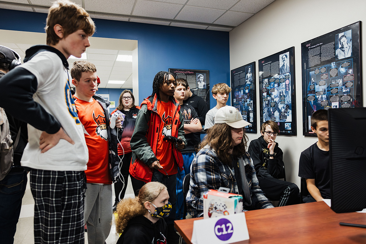 A group of students is gathered around a computer, looking at displays on the wall that feature various images and text. The setting appears to be an educational environment.