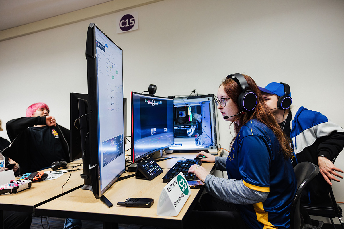 A person wearing a blue esports jersey and headphones is seated at a desk with multiple monitors, a keyboard, and gaming controllers. The setup includes a visible gaming PC. A sign on the desk reads "ESPORTS S3 Streaming."