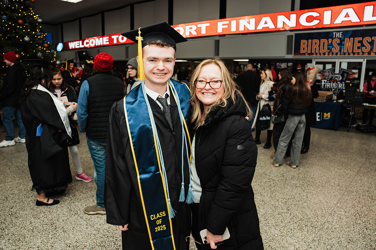 A graduate in a cap and gown with a blue and gold sash stands next to a person in a black coat, smiling in a festive indoor setting. A large sign in the background reads "WELCOME TO SPORT FINANCIAL."