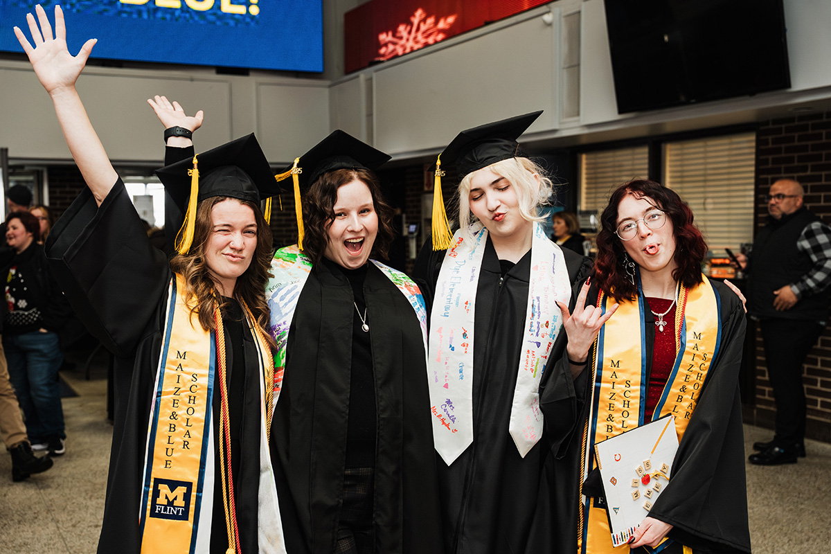 Four graduates in caps and gowns pose together, celebrating with smiles and hand gestures. They wear sashes and hold a diploma. A large screen is visible in the background.