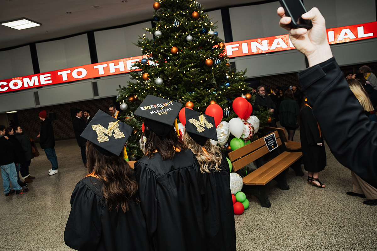 Three graduates in black gowns and decorated caps stand in front of a Christmas tree, with a festive backdrop and balloons.