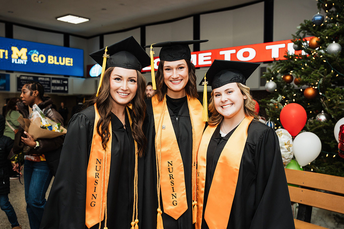 Three graduates in black gowns and caps stand together, wearing orange sashes that read "NURSING." In the background, there is a festive setting with a Christmas tree and colorful decorations.