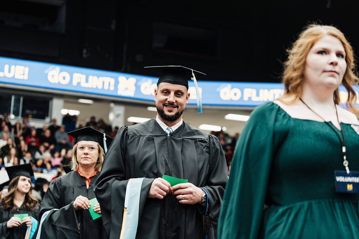 A group of graduates in black caps and gowns are walking in a ceremony, holding green folders. A volunteer in a green dress is also present. A crowd is visible in the background, with a banner that reads "GO FLINT!" above.