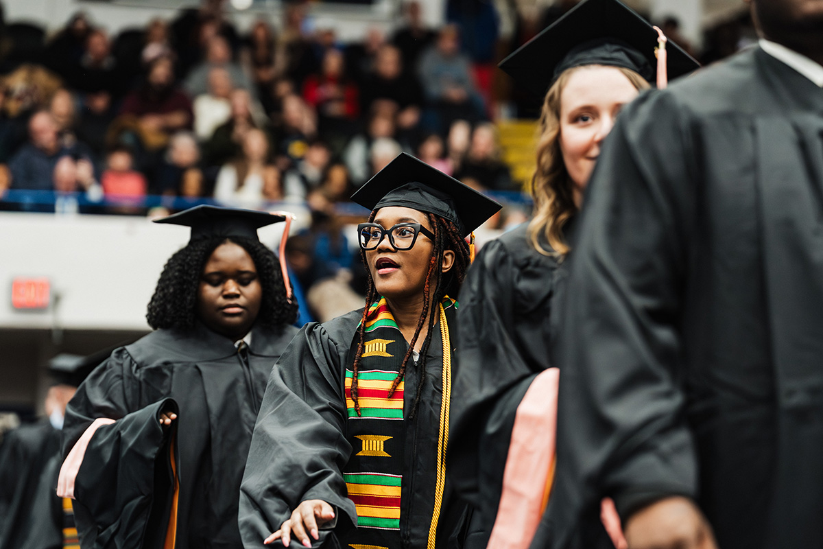 A group of graduates in black caps and gowns walking in a ceremony, with one wearing a colorful kente stole.