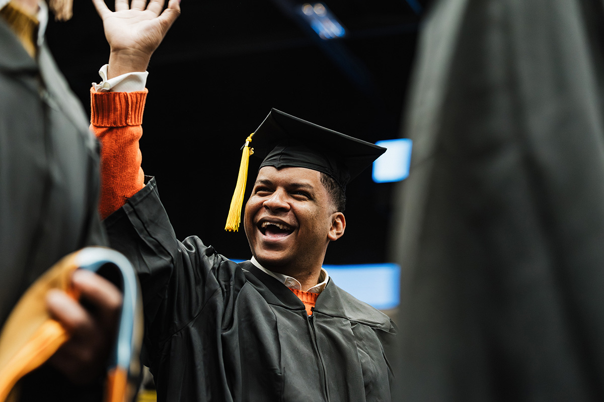 A graduate in a black gown and cap with a yellow tassel raises their hand, surrounded by other graduates.