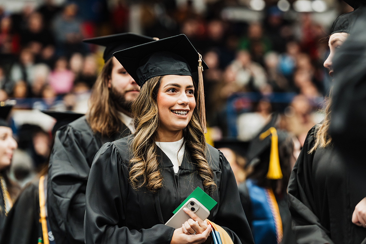 A group of graduates in black caps and gowns, with one holding a phone and a green folder, in a crowded auditorium.