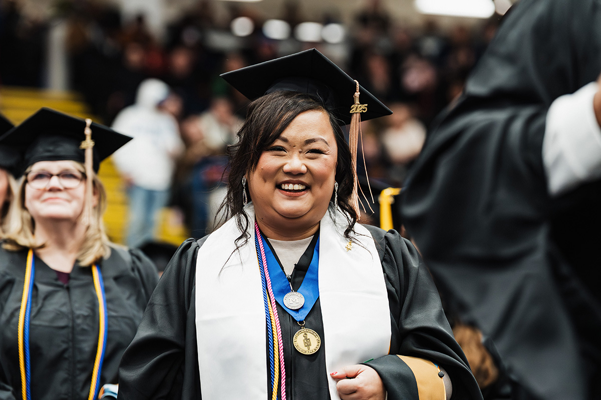 A graduate wearing a black cap and gown, with a white stole and multiple honor cords, stands among fellow graduates in a crowded venue.