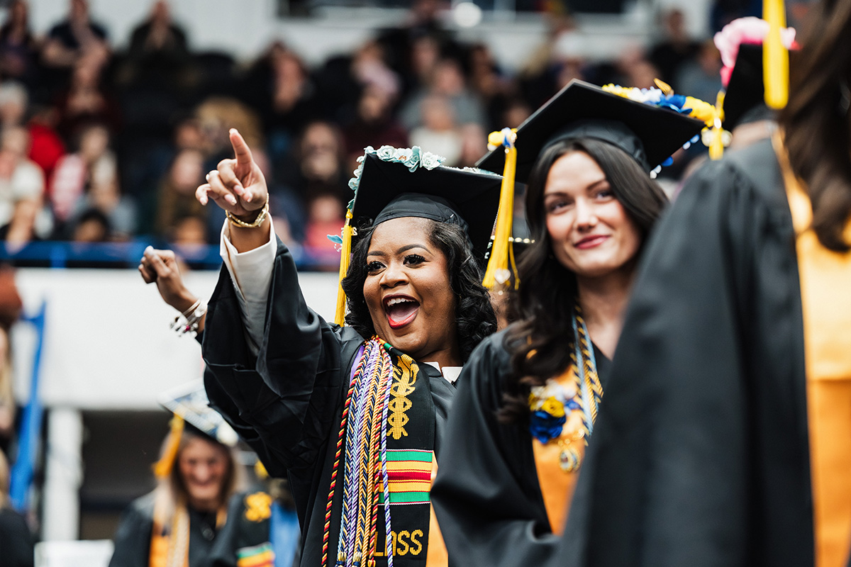 A group of graduates in caps and gowns, one graduate is pointing excitedly while wearing a colorful stole.