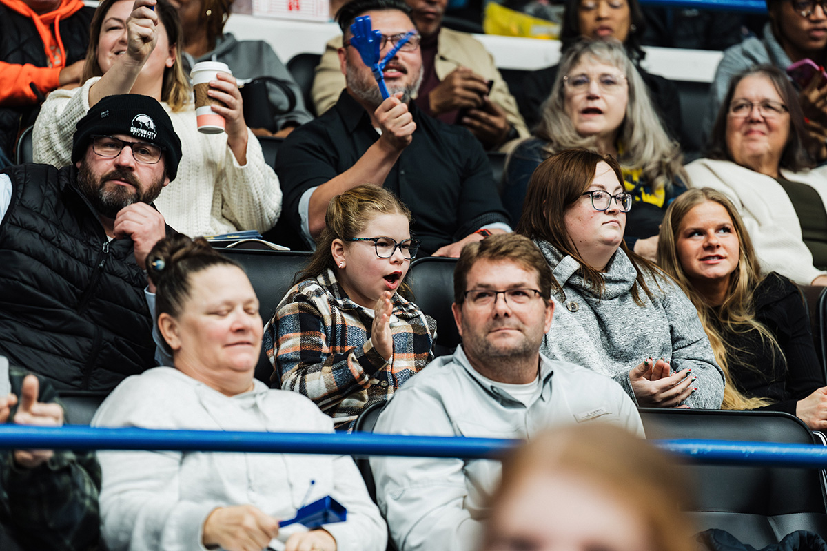 A group of people sitting in a stadium, some holding drinks and others clapping, with a focus on a child in a plaid shirt.