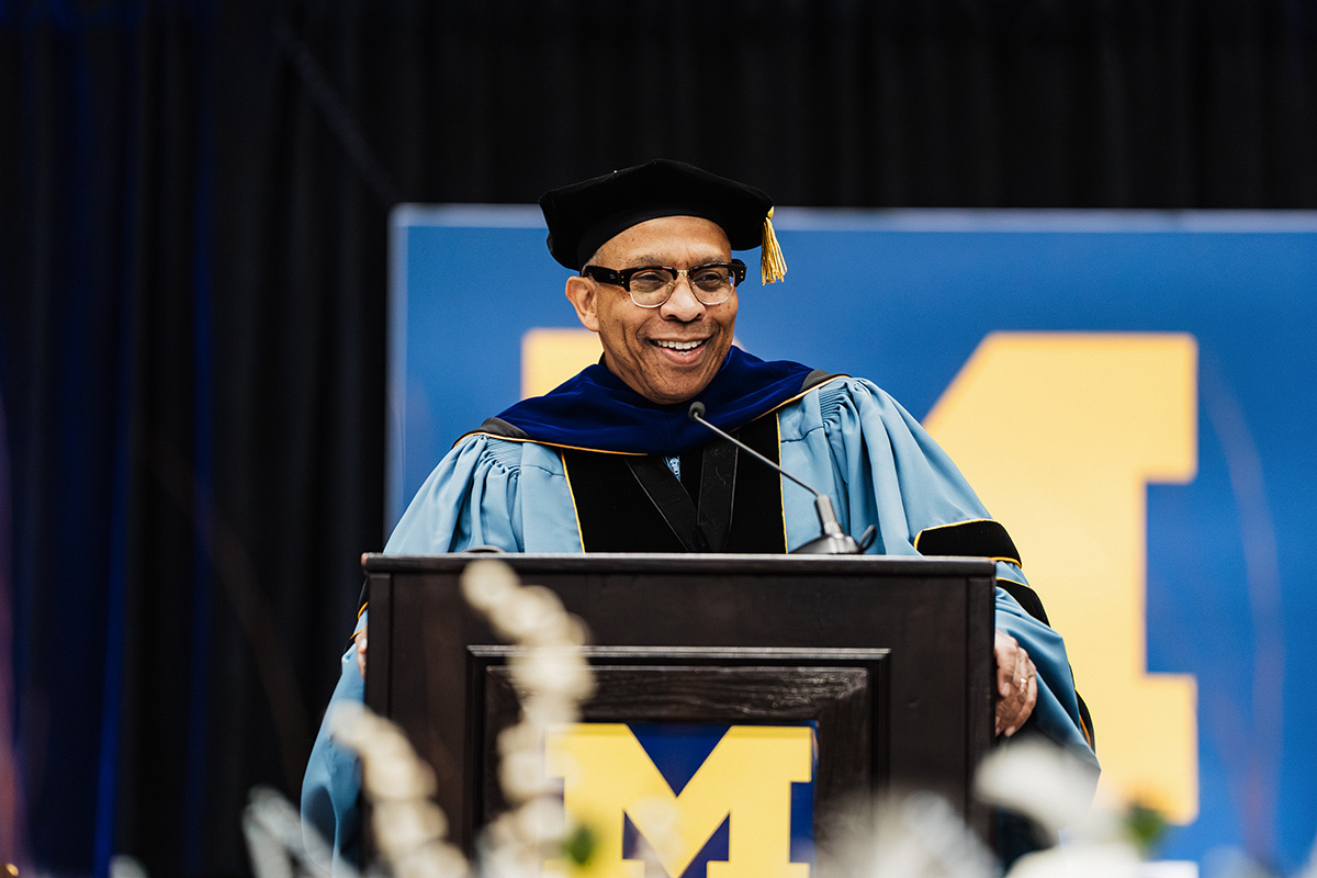 A person in academic regalia stands behind a podium, speaking at a graduation ceremony, with a large "M" logo in the background.