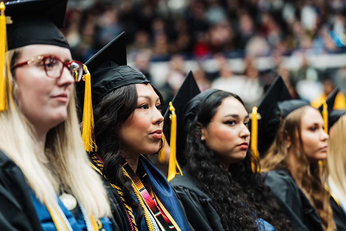 A group of graduates wearing black caps and gowns with yellow tassels, seated in a crowded auditorium during a graduation ceremony.