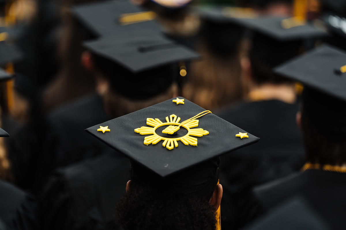 A close-up of a graduation cap decorated with a yellow sunburst design and small stars, surrounded by other black graduation caps.