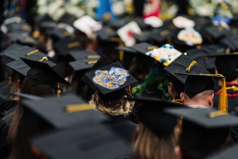 A crowd of graduates wearing black caps and gowns, with one cap featuring a blue and gold WOlverine design.