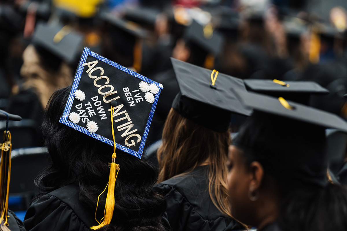 A graduation cap decorated with the text "I'VE BEEN ACCOUNTING DOWN THE DAYS" and adorned with white flowers, set against a background of other graduates.
