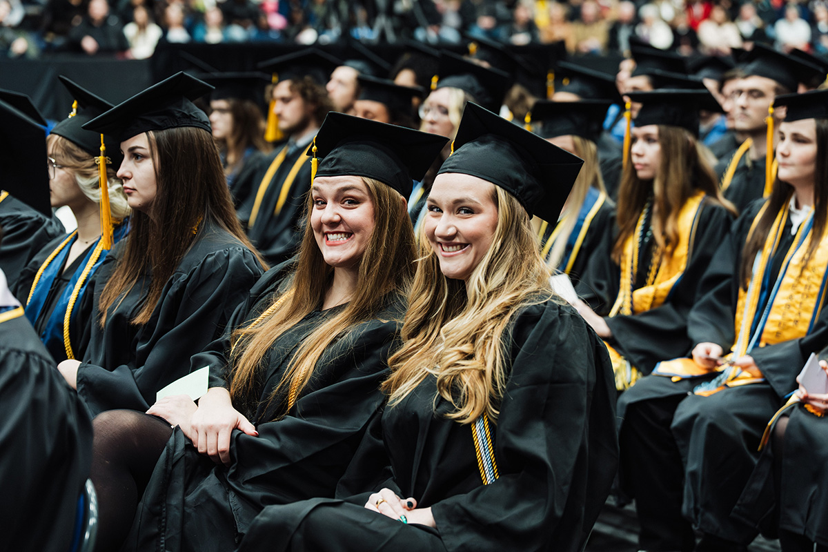 A group of graduates in black caps and gowns, some wearing honor cords, seated in a large auditorium during a graduation ceremony.