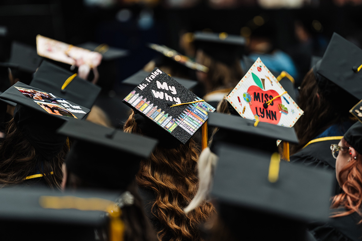 A close-up of graduation caps in a crowd, featuring decorated caps with messages like "My Why" and "Miss Lynn."