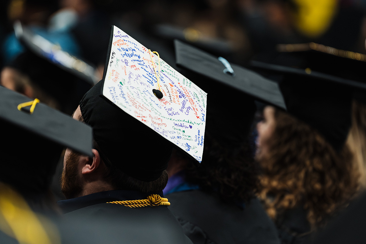 A close-up of graduation caps in a crowd, focusing on one cap decorated with colorful messages and signatures on the top.