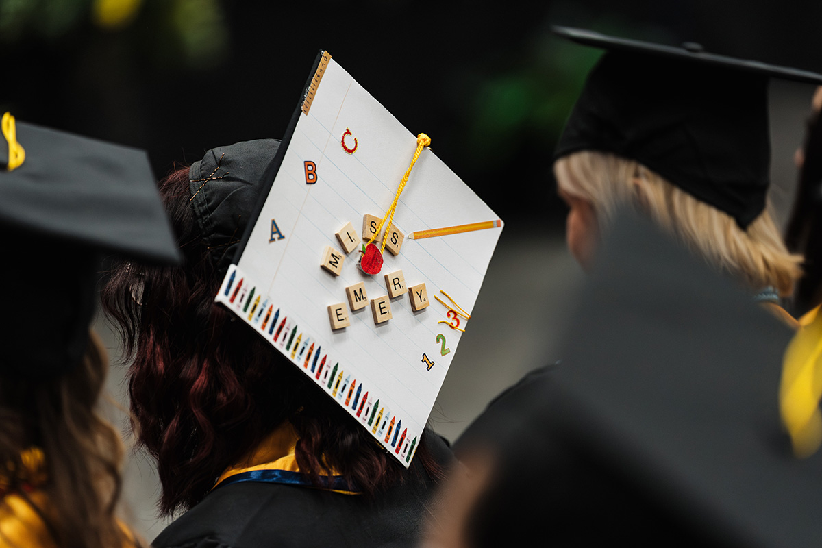 A graduation cap decorated with a design resembling a notebook, featuring the words "MISS EMERY" made from letter tiles, along with a pencil and colorful crayon illustrations.