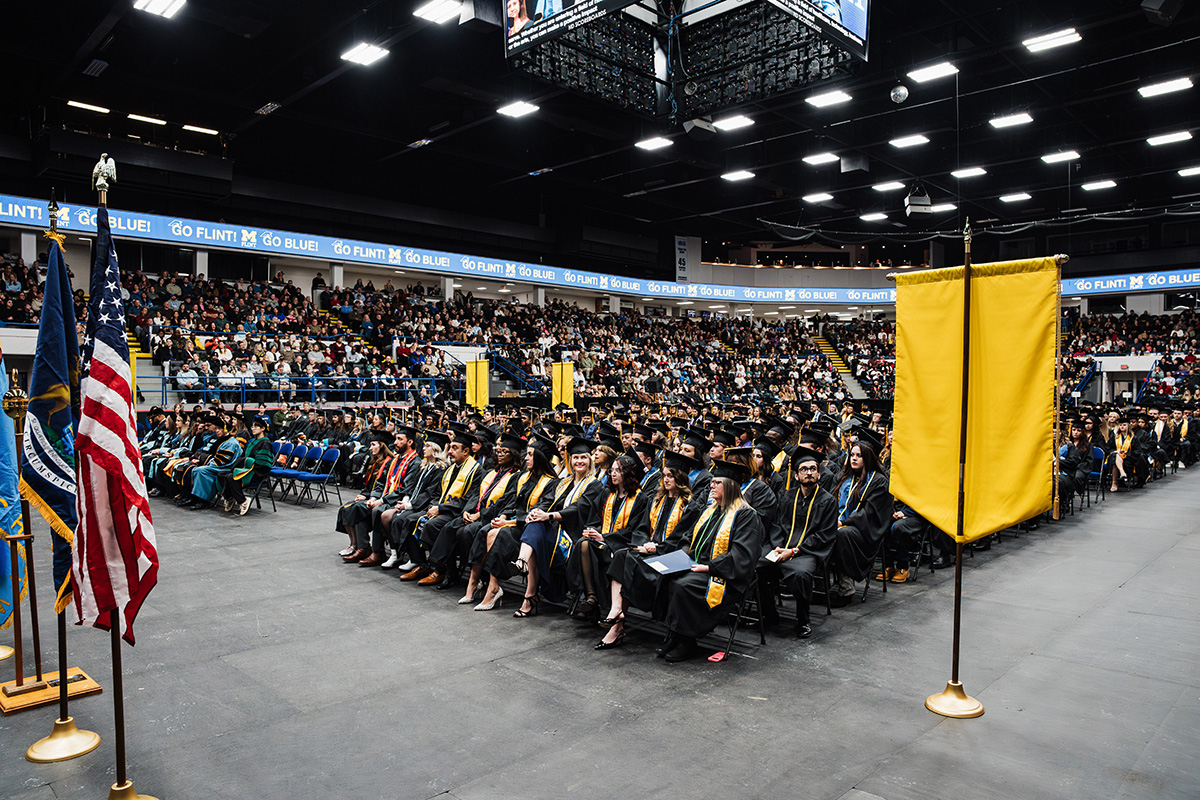A graduation ceremony in a large indoor arena, with graduates in caps and gowns seated in rows. Flags are displayed in the foreground, and a large audience is visible in the background.