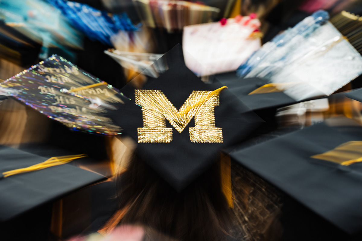 A blurred image of graduation caps, with one prominently displaying a large, glittery gold letter "M" on a black background.