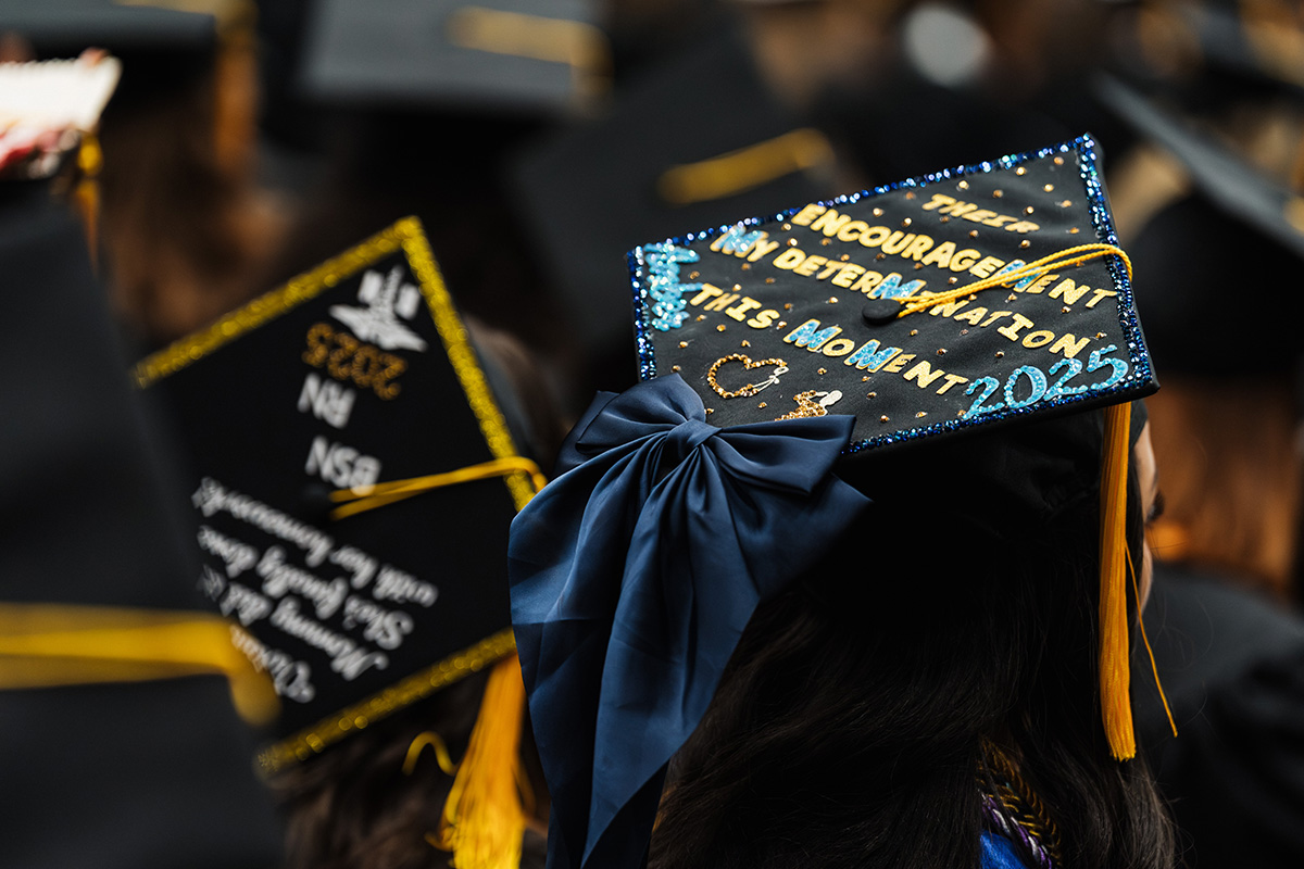 A decorated graduation cap featuring the text "Their encouragement, my determination, this moment 2025" in colorful letters, with a blue bow attached.